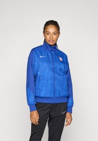 Plava jakna s patentnim zatvaračem i uzorkovanim teksturama, visoki ovratnik i elastični manžeti. Ima Nike logo i grb momčadi na prsima. Uparena je s crnim hlačama.