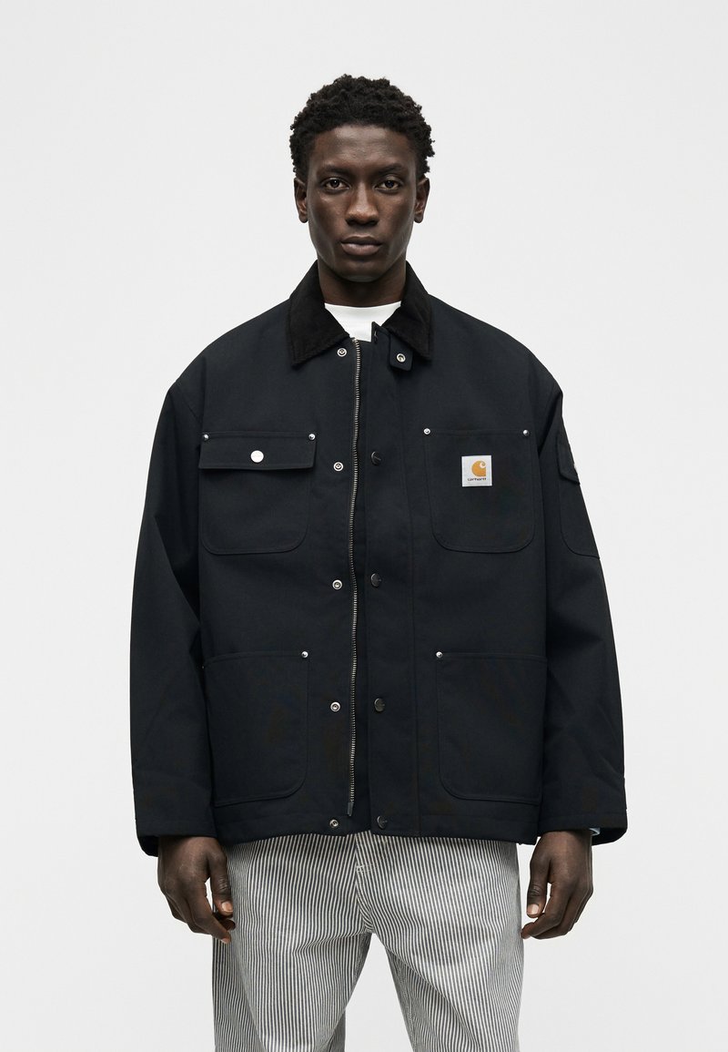 Young man wearing a black jacket with multiple pockets and a Carhartt logo, paired with light gray and white striped pants.