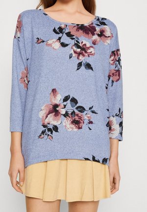 Haut à manches longues bleu clair avec un motif floral en rose et accents foncés, associé à une jupe plissée jaune. Texture en tricot douce.