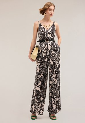 Jumpsuit in bianco e nero con motivo paisley, gamba larga, scollatura a V, cintura nera e dettagli in metallo dorato. La modella porta una borsa dorata e indossa tacchi verdi.