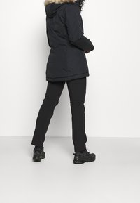 Person mit schwarzer Winterjacke mit Pelzkapuze, schwarzen Hosen und schwarzen Sportschuhen, der mit dem Rücken zu einer weißen Hintergrund steht.