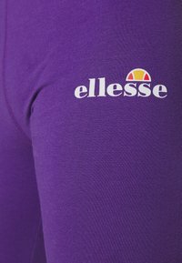 Fialová tkanina s detailním záběrem na logo "ellesse" v bílé barvě, s barevným poloobloukem nad textem.