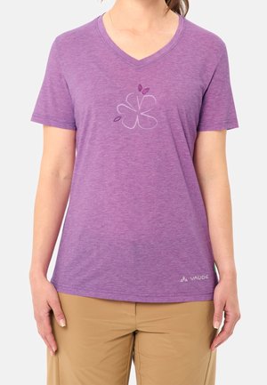 Mujer que lleva una camiseta morada con cuello en V con un diseño minimalista de flores y pantalones beige sobre un fondo liso.