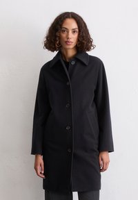 Frau mit lockigem Haar, die einen schwarzen Knopfmantel mit Kragen trägt und vor einer schlichten weißen Wand steht.