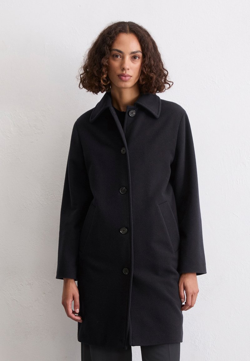 Frau mit lockigem Haar, die einen schwarzen Knopfmantel mit Kragen trägt und vor einer schlichten weißen Wand steht.