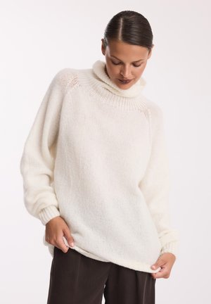 Biały, oversize'owy sweter z golfem wykonany z miękkiego, teksturowanego materiału, z prążkowanymi mankietami i dołem, w połączeniu z ciemnymi, luźnymi spodniami.