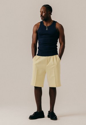 Man met gevlochten haar, gekleed in een zwart mouwloos shirt, beige knielange shorts, zwarte schoenen en gouden kettingen, staat met handen in zijn zakken.