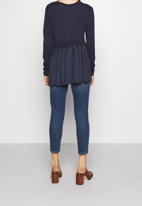 Námořnicky modrý top s dlouhým rukávem a plisovanými zády, doplněný úzkými tmavými džínami a hnědými sandály na podpatku, zvýrazňující vrstevnatou texturu.