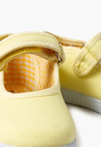 Pantofi sport galbeni din pânză, cu curea ajustabilă, talpă albă și căptușeală cu model în carouri. Caracteristici includ o textură moale și un design cu vârful rotunjit.