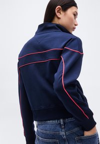 Γυναίκα με σκουρόχρωμα μαλλιά που φοράει μπλε μπουφάν με ροζ λεπτομέρειες στα μανίκια και την πλάτη, συνδυασμένο με μπλε τζιν παντελόνι, γυρισμένη προς τα πίσω.