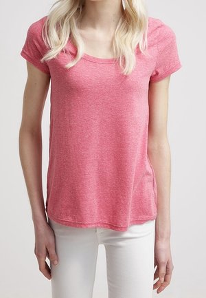 Vrouw draagt een roze T-shirt met korte mouwen en ronde hals en witte broek, staat met ontspannen armen langs haar lichaam.