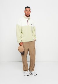 Fleece pullover in light green and white, featuring a half-zip collar and maroon detailing. Beige cargo pants and a cap held in hand.