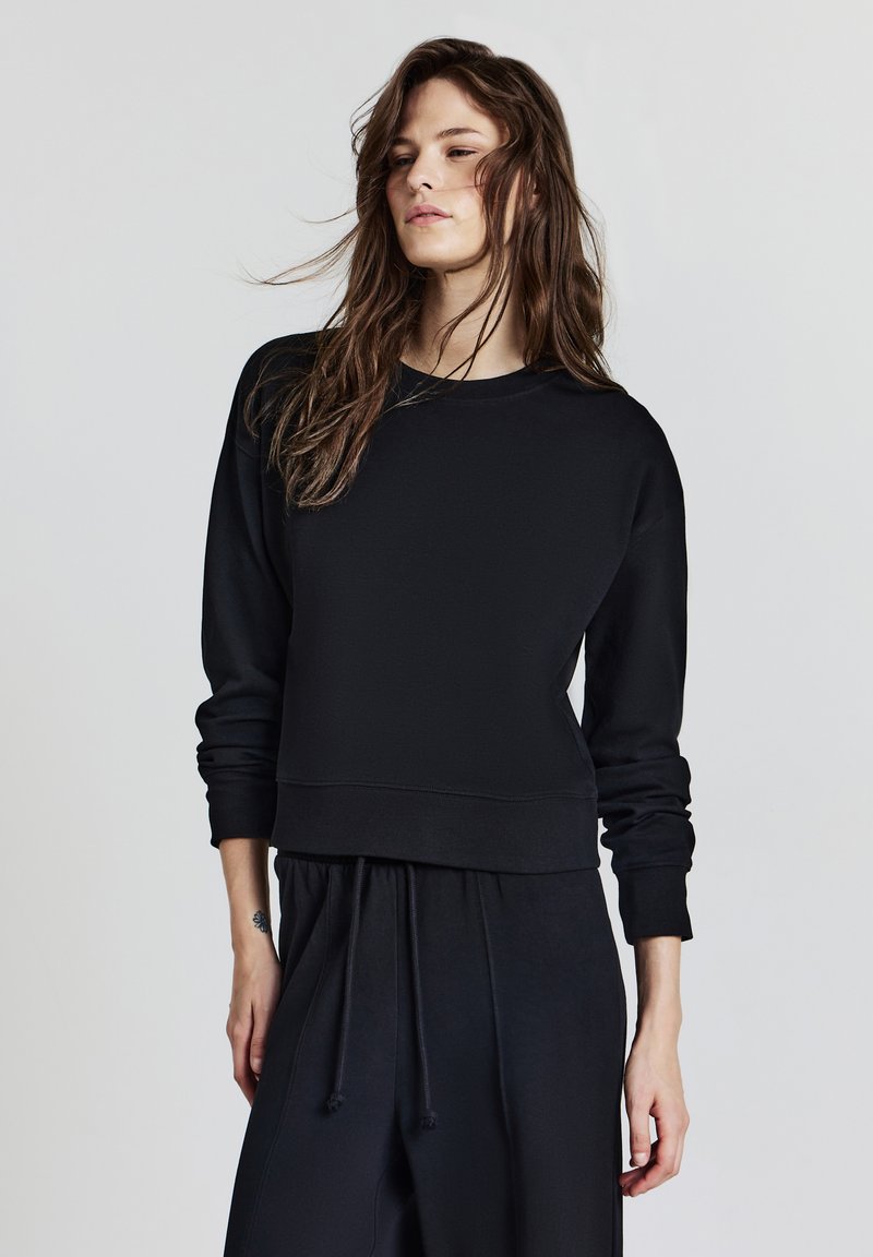 Giovane donna con lunghi capelli castani che indossa una felpa nera e pantaloni neri con cordini, in piedi contro uno sfondo chiaro e semplice.