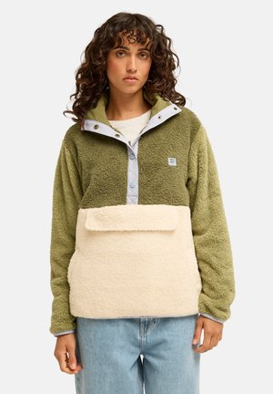 Femme aux cheveux bouclés portant un pull polaire vert et beige avec bouton-pression à moitié et poche avant, associé à un jean bleu clair.