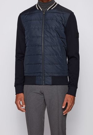 Homme portant une veste matelassée bleu marine avec des manches noires, une fermeture éclair à l'avant, un col rayé et un pantalon gris.