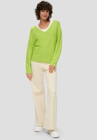 Sweter w kolorze limonkowym z dekoltem w serek i teksturowanym splotem, zestawiony z kremowymi spodniami dzwonami i białymi trampkami na platformie. Model pozujący na neutralnym tle.