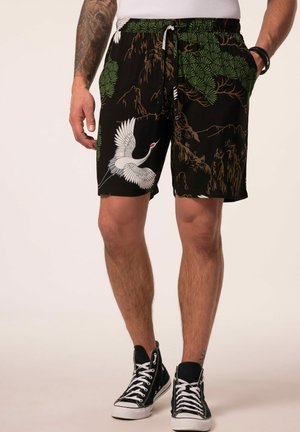 Man wearing black drawstring shorts with green foliage and white crane print, black high-top sneakers, a white shirt, and tattooed arm.