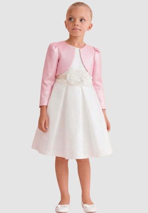 Ragazza giovane che indossa un vestito bianco con dettaglio floreale in vita e una giacca bolero rosa, in piedi con un'espressione neutra.