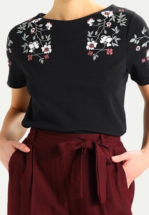 Czarny top z krótkim rękawem z białym i czerwonym haftem kwiatowym, noszony z wysokim stanem bordowymi spodniami przewiązanymi paskiem z tkaniny.