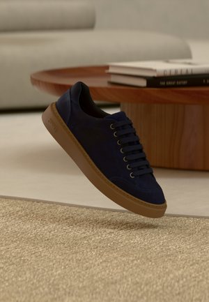 Dunkelblauer Wildleder-Sneaker mit brauner Sohle schwebt über beigem gewebtem Teppich, im Hintergrund ein runder Holztisch und gestapelte Bücher.