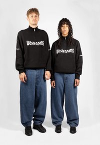 Melni pārāk lieli zipa džemperi ar "Wastedparts" logo, apvienoti ar brīviem zilo denim džinsiem un melnām kurpēm.