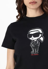 Juodas medvilninis marškinėlis su animaciniu personažu, dėvinčiu akinius nuo saulės ir kostiumą, o apačioje raudonai įrašyta "KARL LAGERFELD".