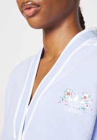 Blue terry cloth garment with a white trim, featuring floral embroidery and an Adidas logo on the left side. Soft texture and relaxed fit.