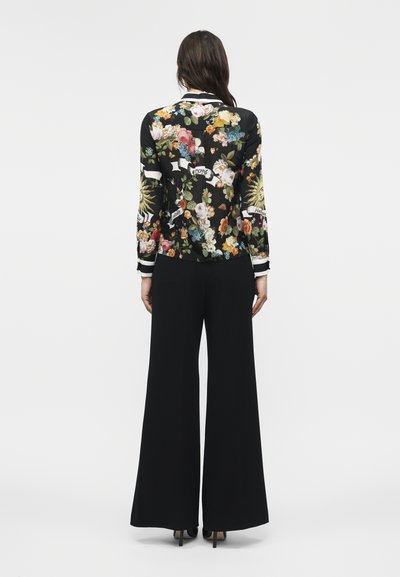 Γυναίκα που φοράει μαύρο παντελόνι με φαρδιά μπατζάκια και μαύρο μακρυμάνικο τοπ με πολύχρωμα λουλούδια και λευκές διακοσμητικές ρίγες, στέκεται γυρισμένη πλάτη.