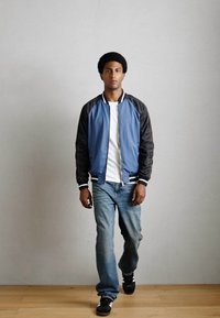 Blaue und schwarze Bomberjacke mit weißen Streifen, kombiniert mit einem weißen T-Shirt und blauen Jeans. Das Modell trägt schwarze Sneaker. Neutrale Hintergrund.