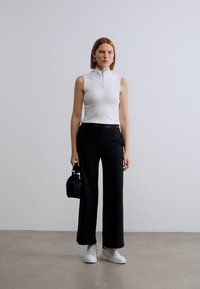 Λευκό αμάνικο τοπ με φερμουάρ, μαύροι φαρδιοί παντελόνες, λευκά αθλητικά παπούτσια και μια μικρή μαύρη τσάντα. Μινιμαλιστικός σχεδιασμός, λείες υφές.