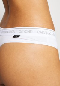 Calvin Klein witte string met een gladde textuur, voorzien van een grijze logo tailleband en een klein zwart label aan de zijkant.