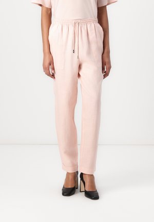 Femme portant un pantalon à cordon rose clair et des talons hauts noirs, debout devant un fond blanc uni.
