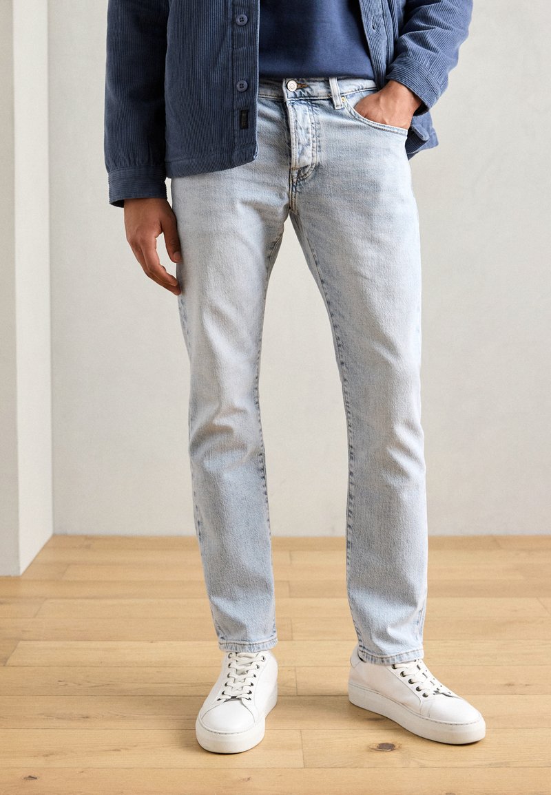 Jeans en denim bleu clair avec une coupe slim, dotés d'un design à cinq poches et d'un léger délavage, associés à des baskets blanches.