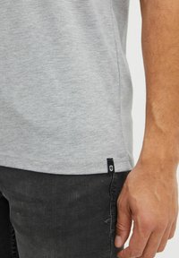 Camiseta gris de manga corta con una textura suave y un dobladillo redondeado, que presenta una pequeña etiqueta de logo negro en la costura lateral, combinada con jeans negros.