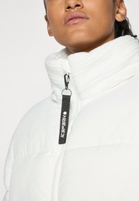 Närbild av en person som bär en vit vadderad jacka med hög krage och ett svart dragflöjt med texten "Icepeak."