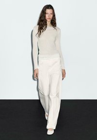 Vrouw die over de catwalk loopt in een getextureerde witte top met lange mouwen, losse witte broek en witte schoenen, tegen een plain achtergrond.