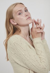 Lys beige strikket sweater med V-udskæring, prydet med et tekstureret mønster og åbne stingdetaljer. Modellen poserer med hænderne foldet nær ansigtet.