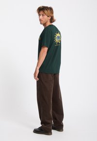 Jeune homme aux cheveux châtain clair portant un t-shirt graphique vert foncé, un pantalon marron ample et des chaussures noires, debout de profil sur un fond blanc.