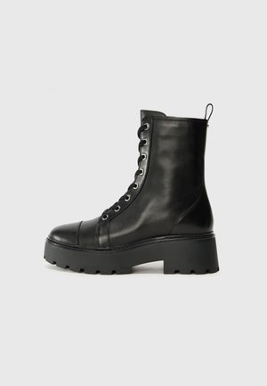 Bota ankle de couro preto com bico redondo, sola de borracha grossa e ilhós de metal. Apresenta um design com cadarços e uma aba de puxar na parte de trás.