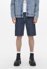 Tummansiniset farkkushortsit suoran leikkauksen omaavina, yhdistettynä vaaleansiiniseen farkkutakkiin ja mustiin nilkkasaappaisiin. Valkoiset perussukat näkyvät.