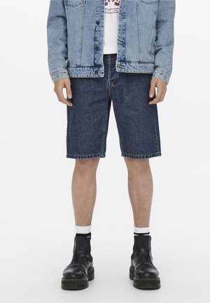 Donkere spijkershorts met een rechte snit, gecombineerd met een lichtblauwe spijkerjack en zwarte enkellaarsjes. Witte crew sokken zichtbaar.