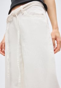 Άτομο που φοράει εκρού παντελόνι με φαρδιά μπατζάκια και υφασμάτινη ζώνη δεμένη στη μέση, σε συνδυασμό με μαύρο τοπ, τα χέρια ακουμπισμένα στα πλάγια.