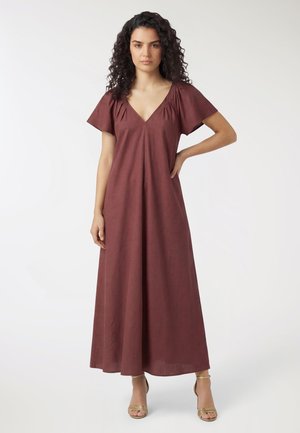 Donna con capelli scuri e ricci che indossa un abito bordeaux ampio, a maniche corte, con scollatura profonda a V, lungo fino alla caviglia, e tacchi dorati con cinturini.