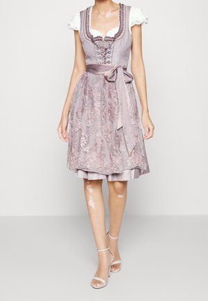 Femme portant une robe dirndl lilas jusqu'aux genoux avec tablier en dentelle florale, nœud en satin à la taille, blouse blanche avec manches en dentelle et sandales à talons beiges.