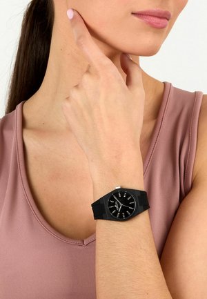 Donna che indossa una maglietta senza maniche color malva con orologio nero al polso, che indica il collo con l'indice.