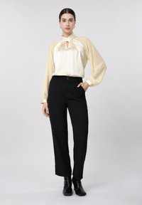 Kvinde stående iført en beige langærmet bluse med halsbind, sorte højtaljede bukser og sorte ankelstøvler mod en ensfarvet baggrund.