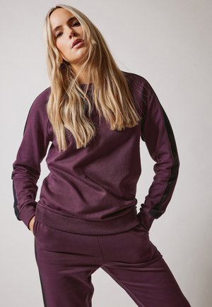 Sudadera púrpura con rayas negras a los lados, con mangas largas y ribete acanalado, combinada con pantalones de chándal púrpuras a juego. Textura de tejido suave.