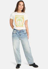 T-shirt graphique blanc avec un design de palmier en vert et orange, associé à un jean délavé, coupe ample et des chaussures noires.