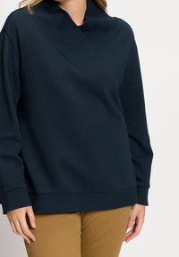 Marinblå pullover med en bred krage och ribbat mönster; avslappnad passform med långa ärmar och rak nederkant, tillsammans med bruna byxor.