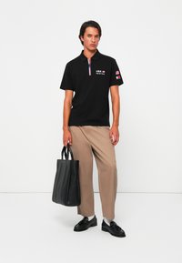 Sort polo skjorte med logo, korte ærmer og to-knaps lukning; beige vide bukser; sort læder taske; sorte sko.
